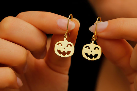 Gold pumpkin-shaped earrings held between fingers against a dark background