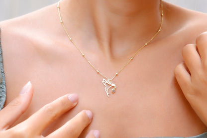A close-up image of a woman's neck wearing a gold-colored necklace with a bird charm and beaded ball chain.