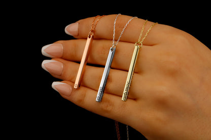 Three necklaces with long bars in silver, gold, and rose gold colors, held between fingers with a dark background.