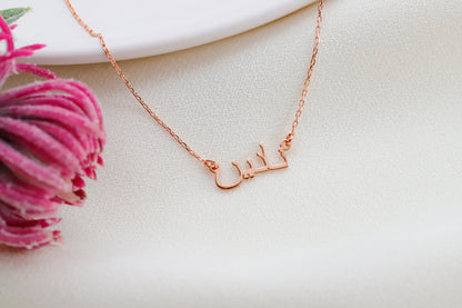 A woman wearing a black tank top showcasing a silver necklace with Arabic script.