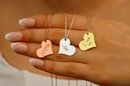 Three heart-shaped necklaces with engraved messages on a hand against a neutral background