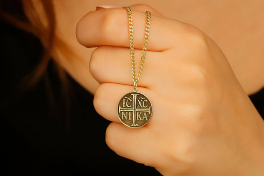 Hand holding a gold necklace with a circular pendant with the Christian Eastern Orthodox cross and the initials IC XC NIKA.