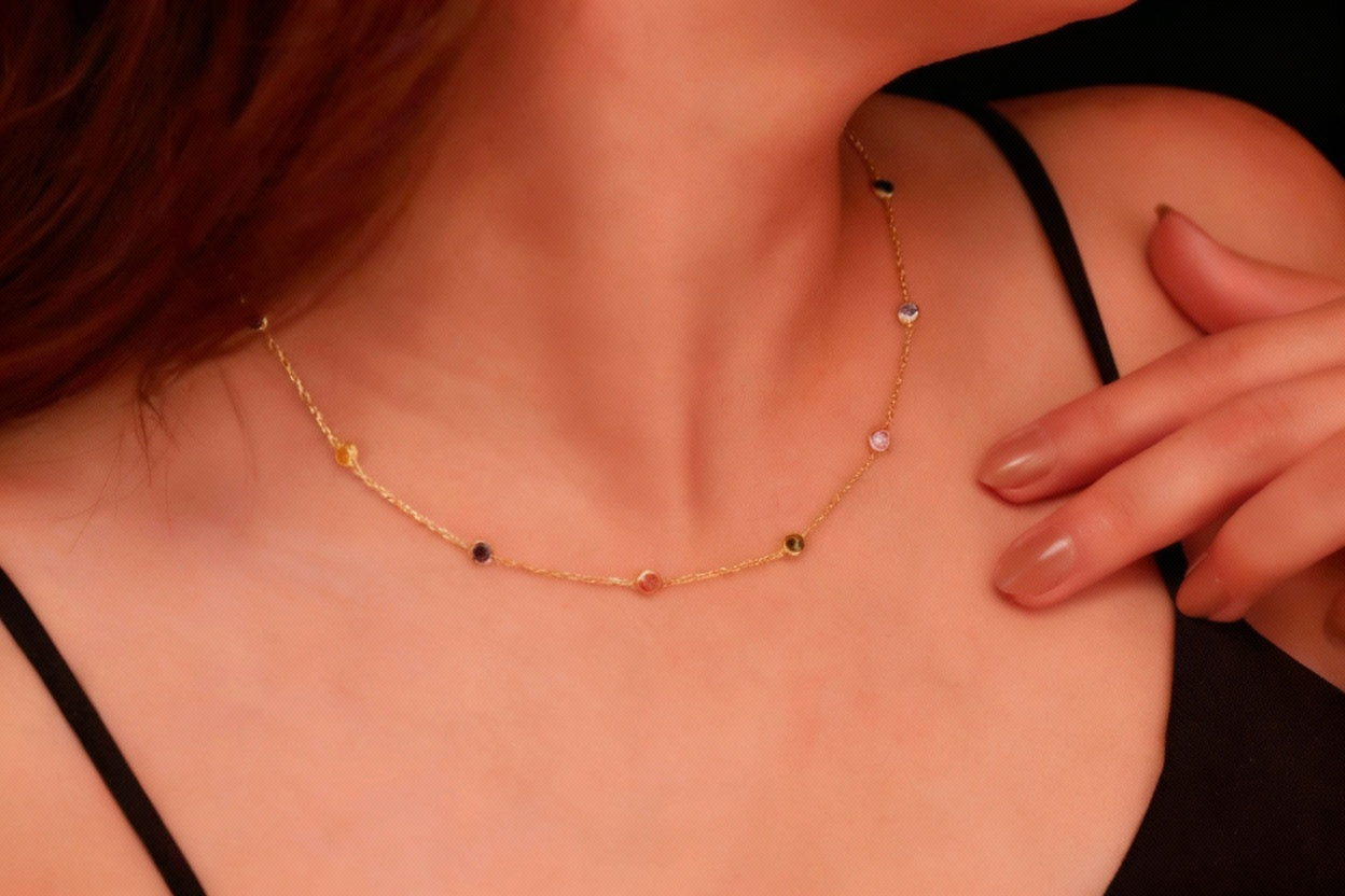 A person wearing a multi-station rainbow necklace with crystal gemstones on a dainty chain, with options in gold and rose gold colors.