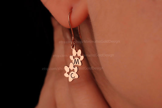 A pair of custom initial letter earrings, with one earring featuring a paw print, engraved with the letter 'M', made of sterling silver, displayed on a person's ear.