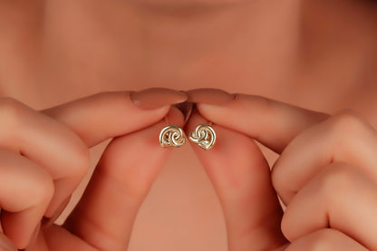 A pair of small dainty stud earrings with a love knot design, held between fingers, showcasing the jewelry's delicate and minimalist design.