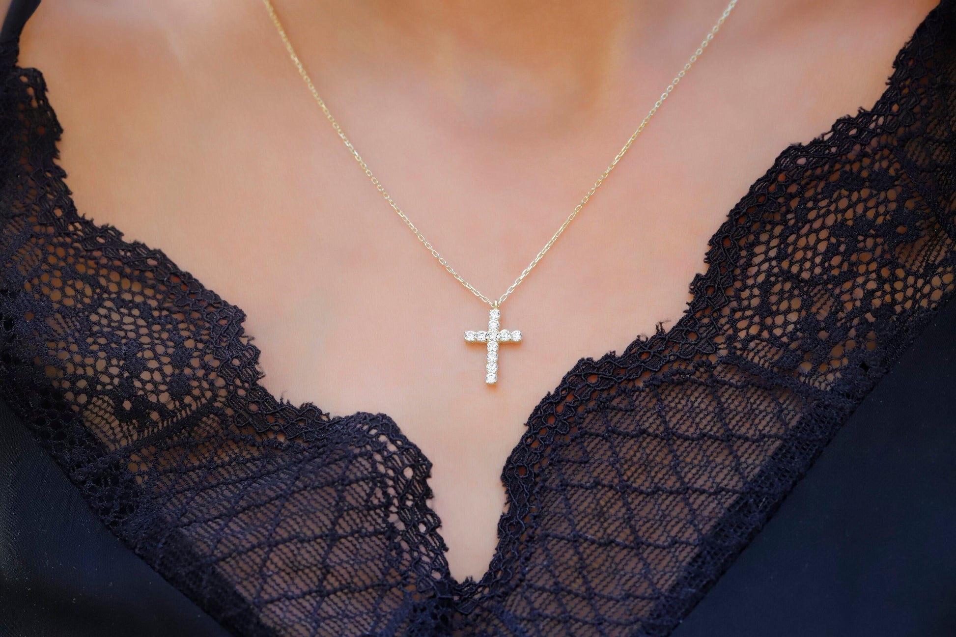 A close-up image of a person wearing a 14K gold diamond cross necklace with a delicate chain, displayed against a lacy black background.