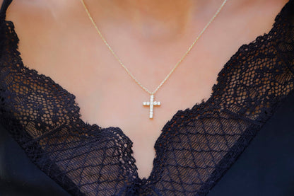 A close-up image of a person wearing a 14K gold diamond cross necklace with a delicate chain, displayed against a lacy black background.