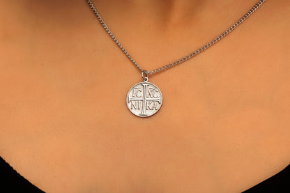 A close-up image of a person wearing a necklace with a round rose gold charm engraved with the Christian Eastern Orthodox cross and the initials IC XC NIKA.