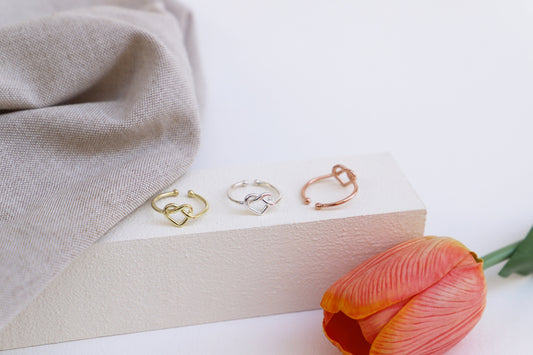 Three adjustable gold heart knot rings on a beige wooden stand, with a neutral-toned fabric and a red flower nearby.