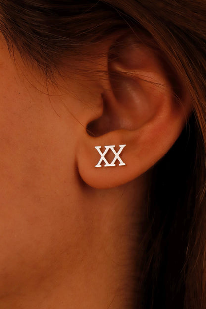 A close-up image of a woman's ear wearing a gold stud earring with the Roman numeral 'XX' displayed.