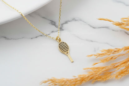 Gold necklace with a tennis racket pendant on a marble surface with dried grass.