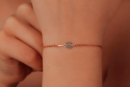 Rose gold bracelet with tennis racket design on a wrist against a warm-toned background
