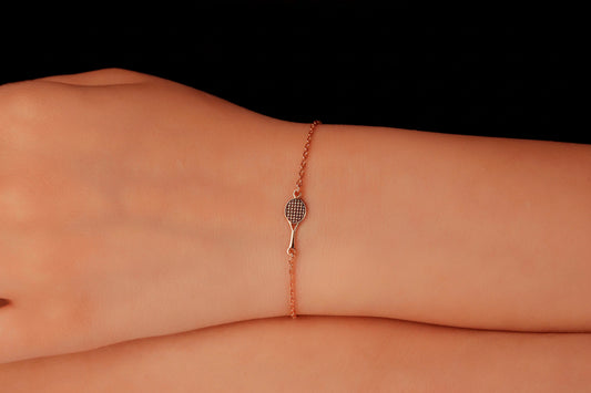 Rose gold bracelet with tennis racket charm on a wrist against a black background