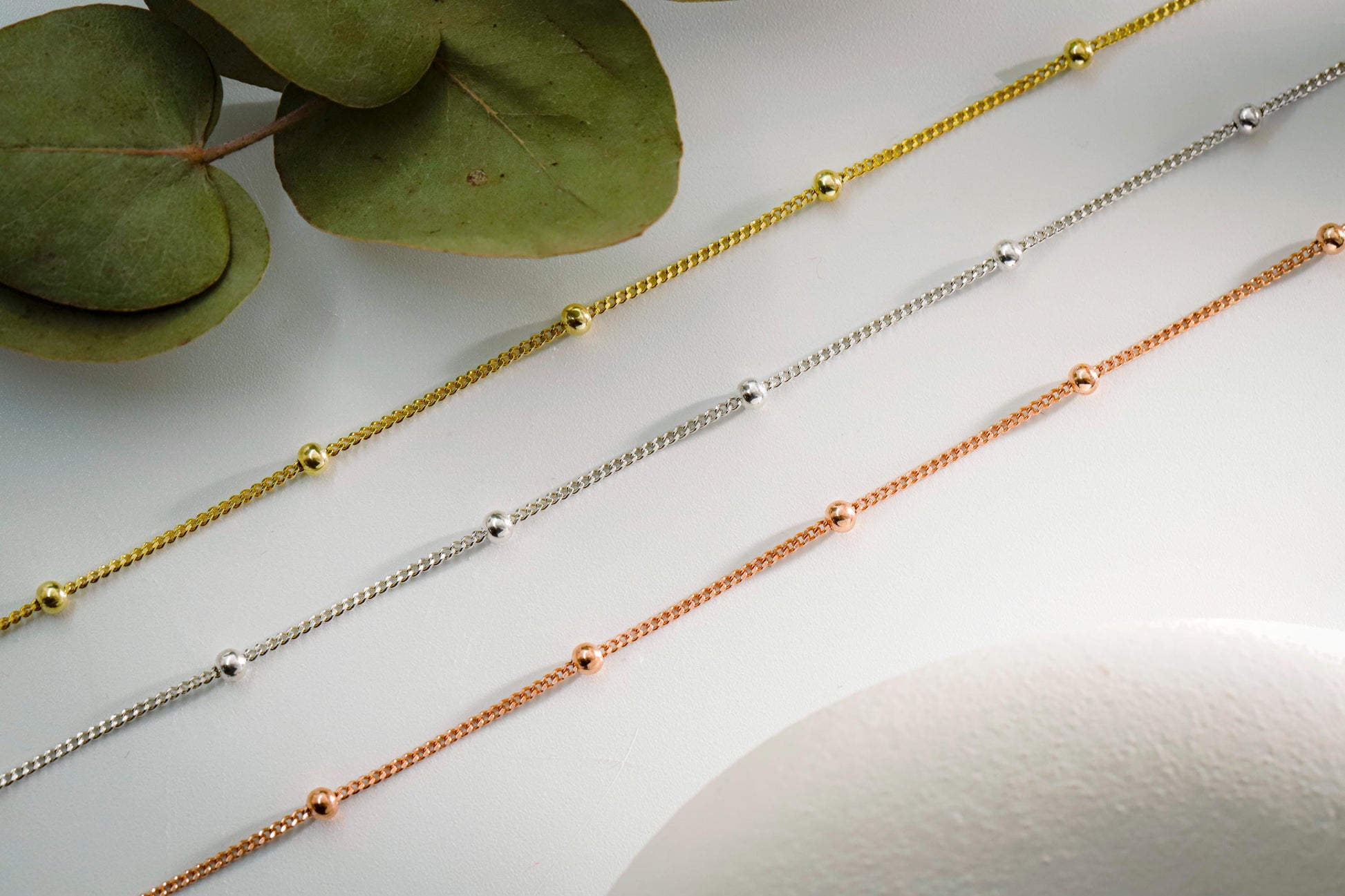 Three gold chain necklaces on a light surface with green leaves in the corner.