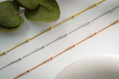 Three gold chain necklaces on a light surface with green leaves in the corner.