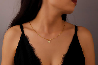 A close-up image of a rose gold monogram necklace with a tiny initial pendant on a box chain, worn by a person.