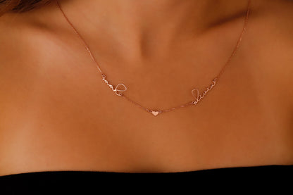 A close-up image of a custom necklace featuring two names in cursive handwriting and a small heart charm, displayed on a person's neck.