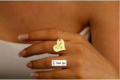 Hand wearing a gold necklace with a heart pendant labeled 'I love you' against a neutral background