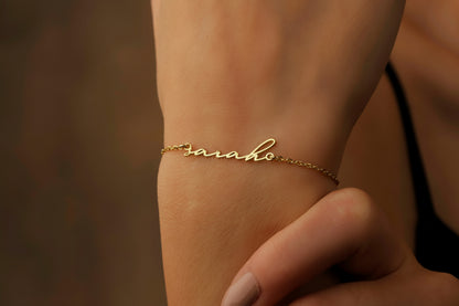 A gold bracelet with the name 'Sarah' written in cursive script on a person's wrist.