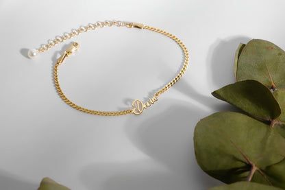 Gold anklet with 'Devin' engraving on a light gray background with green leaves.