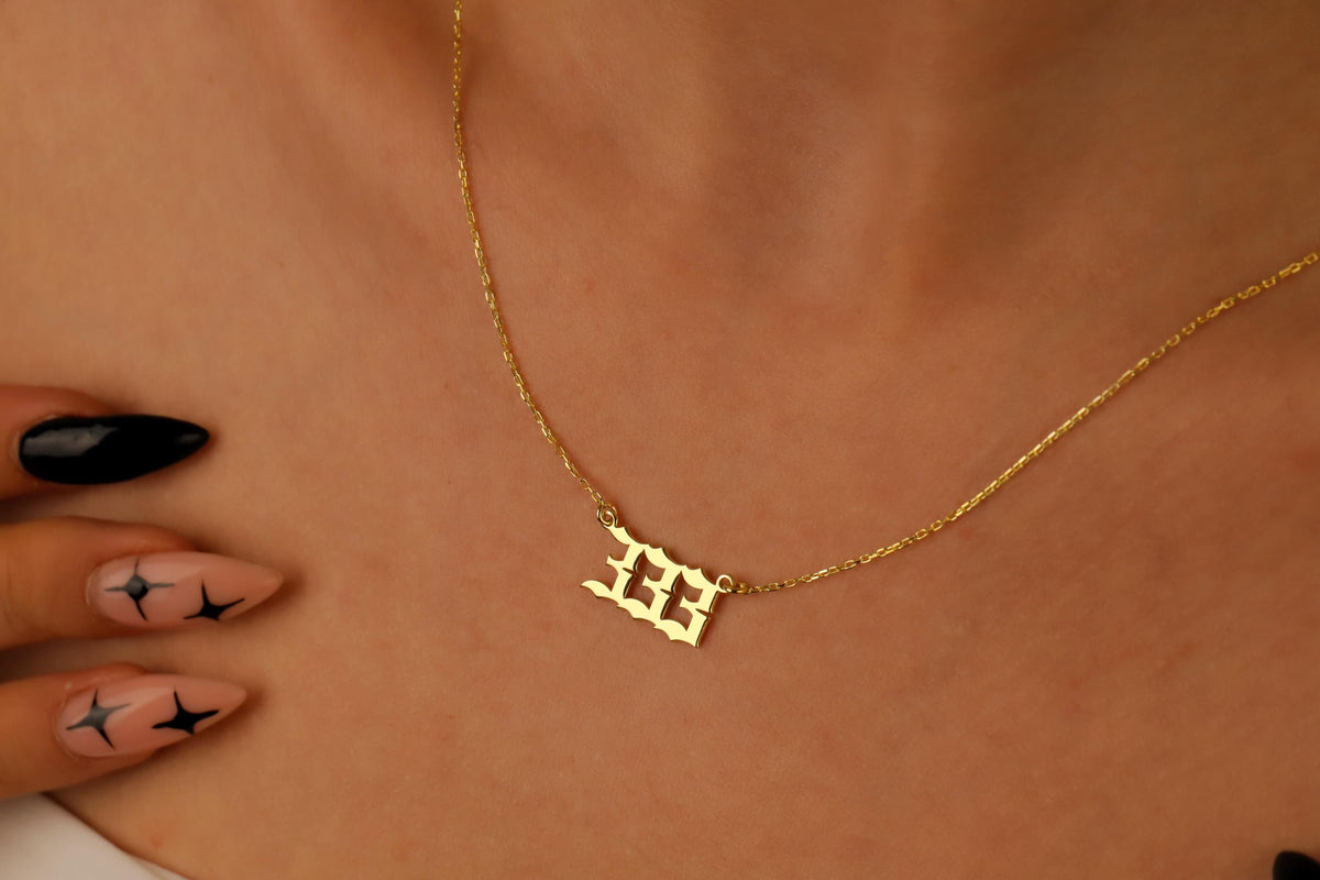 A close-up image of a necklace with the number 333 on a charm, held by a person with red nail polish.