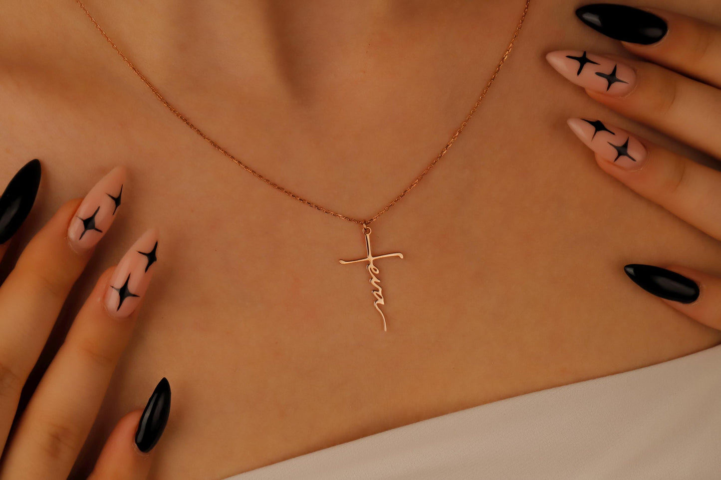 Gold necklace with a cross pendant on a beige surface, with hands wearing black nail polish holding the necklace.