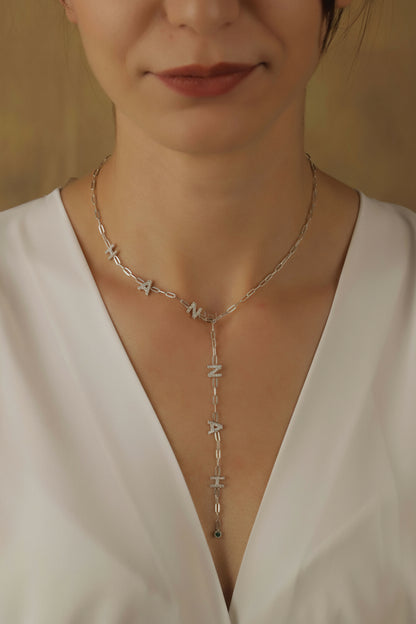 Necklace with lettered pendant worn by a person in a white top against a neutral background