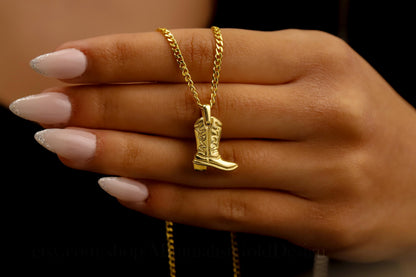 A close-up image of a person's hand holding a gold-colored necklace with a cowboy boot charm attached to a curb chain.