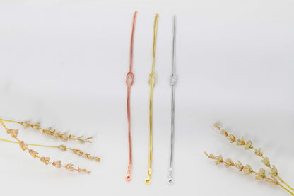 Three love knot bracelets made of 925 sterling silver, displayed in two different finishes, gold and rose gold, alongside decorative dried plants on a white background.