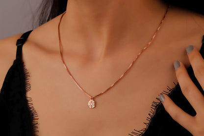 A close-up image of a rose gold monogram necklace with a tiny initial pendant on a box chain, worn by a person.