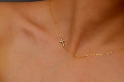 A close-up image of a gold sideways cross necklace with a small CZ stone, displayed on a person's neck.