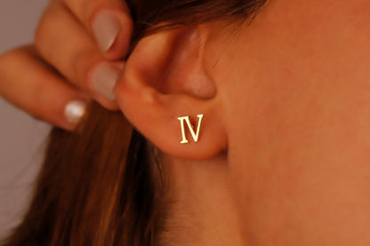 A close-up image of a woman's ear wearing a gold stud earring with the Roman numeral 'IV' displayed.