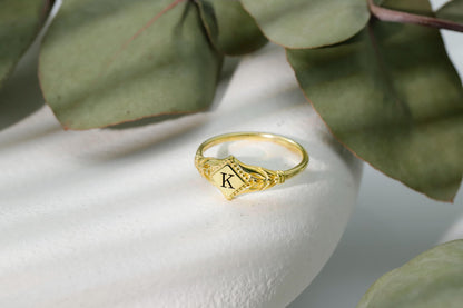 A dainty rhombus-shaped initial ring with a gold-filled design, resting on a white surface with green leaves in the background.