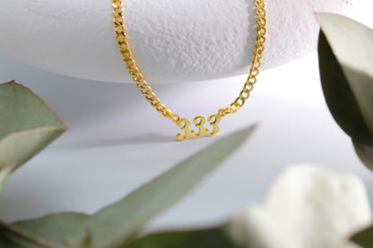 A gold-colored necklace with the numbers '333' displayed, placed on a white surface with green leaves in the background.