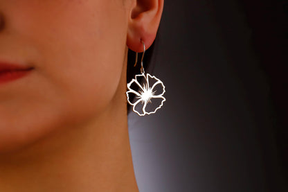 Three birth flower earrings in different colors (gold, silver, rose gold) displayed on a white surface with a green leaf in the background.