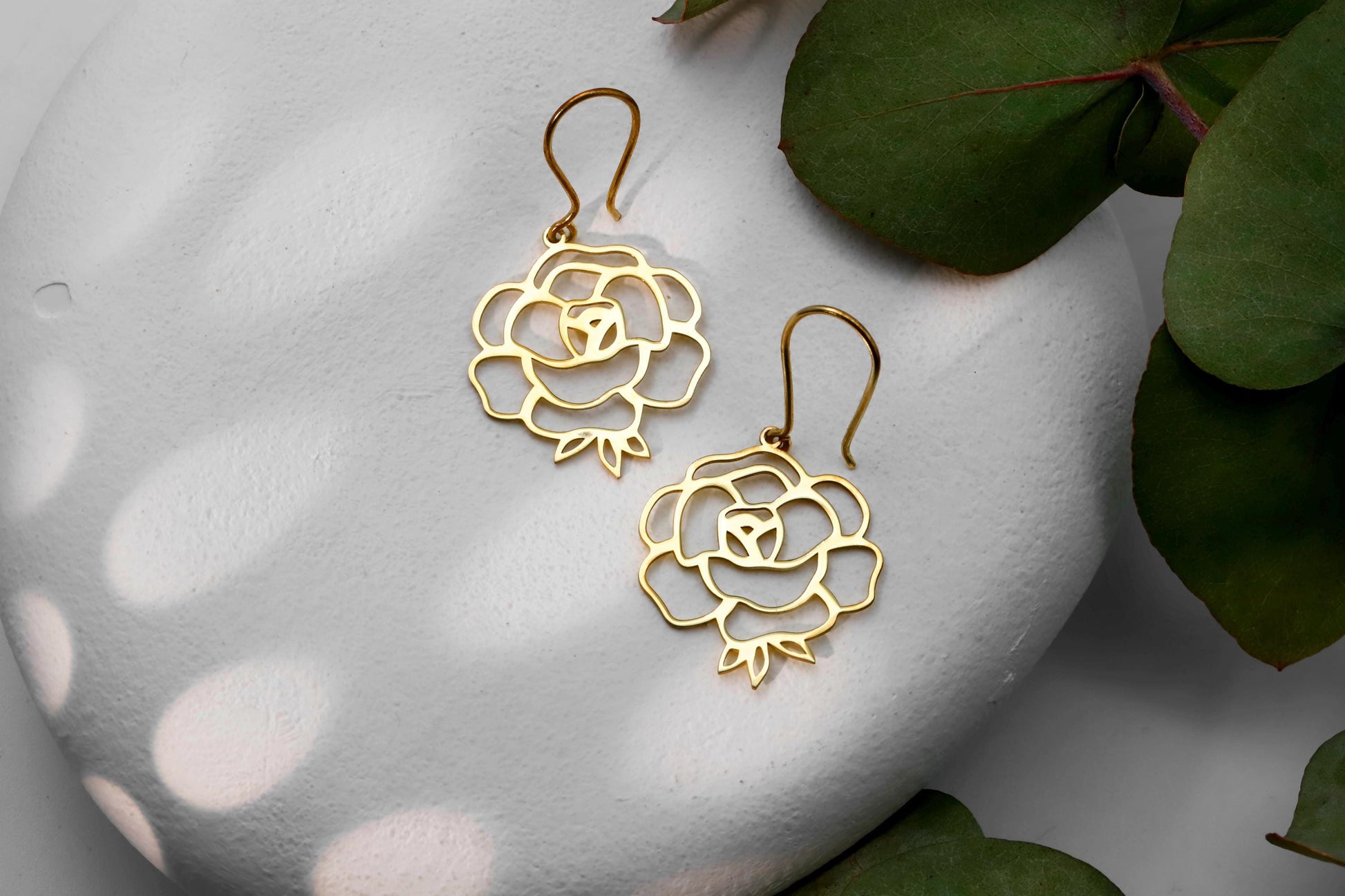 Three birth flower earrings in different colors (gold, silver, rose gold) displayed on a white surface with a green leaf in the background.