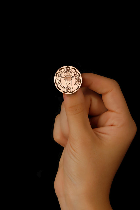 a hand holding rose gold-colored brooch with an engraved university seal 