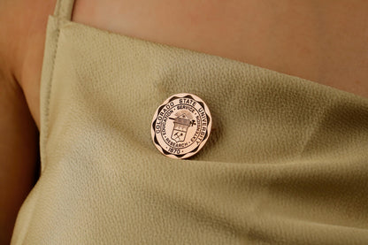 A gold-colored brooch with an engraved university seal placed on a white fabric with a floral pattern.