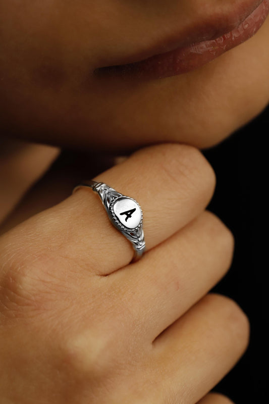 A sterling silver initial ring with the letter 'A' inscribed, placed on a white surface with green leaves in the background.