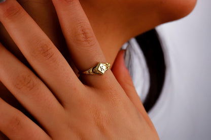 A dainty rhombus-shaped initial ring with a gold-filled design, resting on a white surface with green leaves in the background.