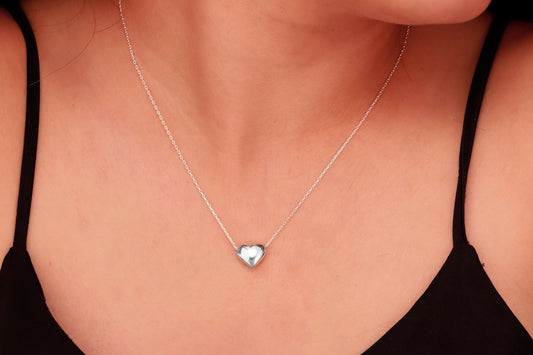 
A silver, heart-shaped pendant hangs from a thin silver chain on a person's neck. A product by SilverGoldandRose.com.