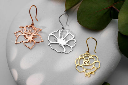 Three birth flower earrings in different colors (gold, silver, rose gold) displayed on a white surface with a green leaf in the background.