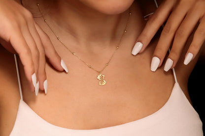 A close-up image of a gold initial necklace with a beaded ball chain, featuring a CZ stone on the initial pendant, worn by a person.