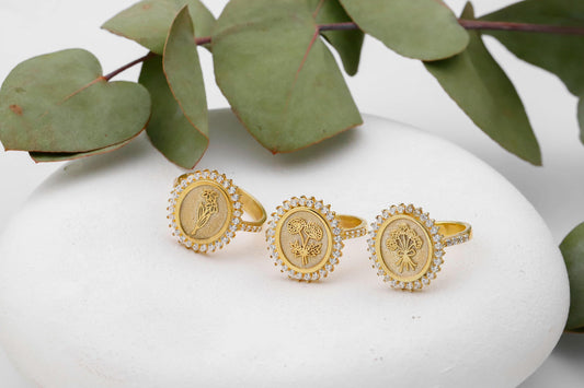 Three gold rings with intricate designs on a white surface with green leaves in the background