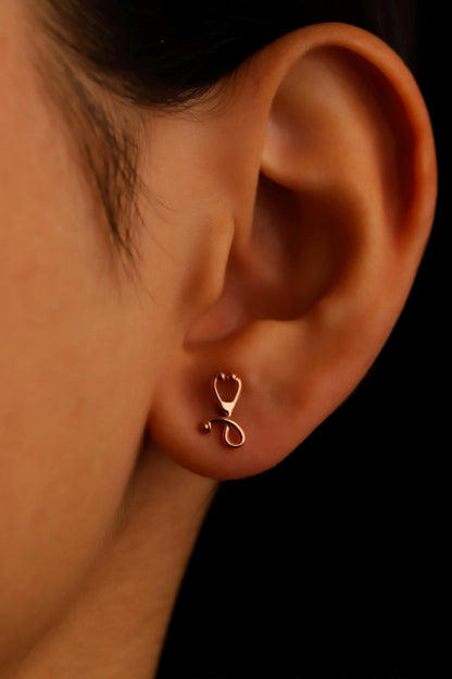 Close-up of an ear wearing a rose gold stethoscope-shaped earring against a black background