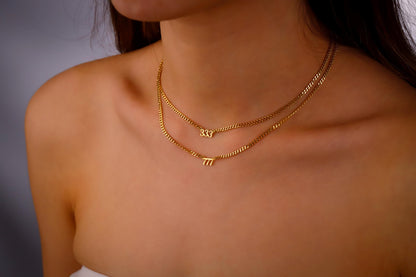 A close-up image of a person wearing a layered gold intertwined necklace with the numbers '833' and '711' on small, dainty chain links.