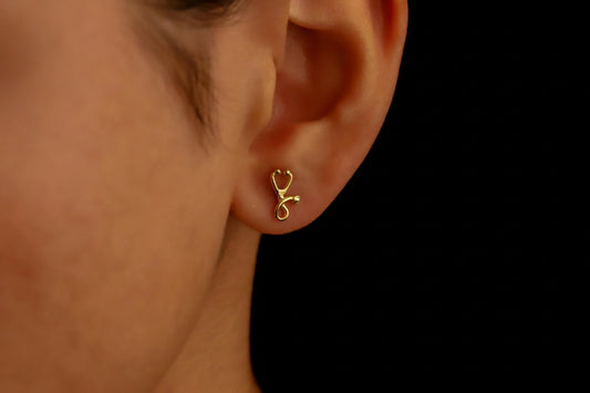 Gold stethoscope-shaped earring worn on a person's ear against a black background