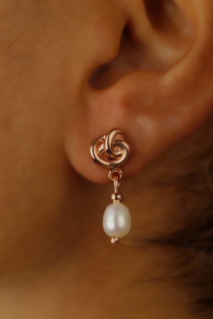 A close-up image of a woman's ear wearing a gold-colored Celtic knot dangle earring with a pearl drop.