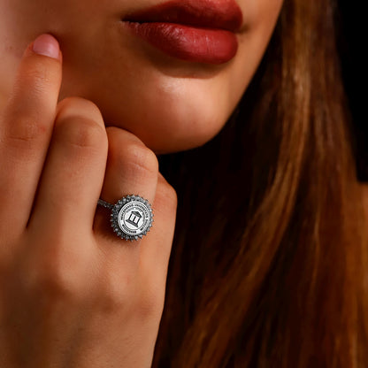 A close-up image of a person's hand showcasing a university ring with a round centerpiece diamond, surrounded by smaller diamonds, and inscriptions on the band.