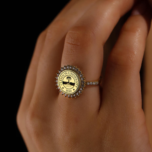 A close-up image of a person's hand wearing a custom university ring with a seal and diamond accents around the band.